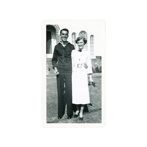 May include: A vintage black and white photograph of a smiling couple. The man is in a navy uniform, and the woman wears a white dress and jacket. They stand on a lawn in front of a building with arched windows.
