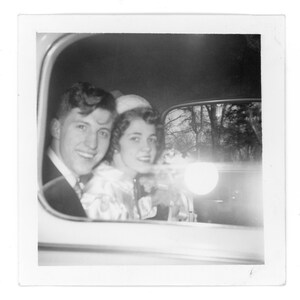 May include: Black and white photograph of a smiling couple in a car. The man is wearing a suit and tie, and the woman is wearing a dress and a hat. The car window frames the couple with a bright light.