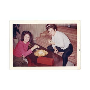 May include: Two women are sitting on a sofa in a living room. One woman is wearing a pink jumper and the other is wearing a white jumper. They are both eating crisps and dip from a bowl on a coffee table.