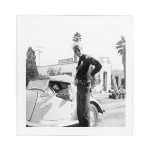 May include: A black and white photograph of a police officer standing next to a vintage car. The officer is wearing a uniform and a hat. The car has a canvas top and is parked in front of a building with the sign "Lawn Mower Shop".