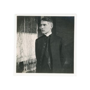 May include: Black and white photograph of a person wearing a dark coat with multiple buttons, a white collar, and a dark vest. The person is standing in front of a building with a brick wall and a window with vines.