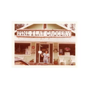 May include: A vintage photograph of a small-town grocery store, "PINE FLAT GROCERY", with a person standing in the doorway. The store has a light green exterior, white trim, and a "Coors Beer" neon sign. A sign for hunting and fishing licenses is visible.