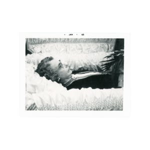 May include: Black and white photograph of a person lying in a casket. The person is wearing glasses and a dark-colored garment. The casket is lined with white fabric. The text "JAN - 65" is in the upper portion of the image.