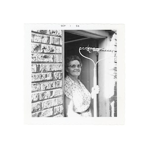 May include: Black and white photograph of a woman in a doorway holding a rake. The woman is wearing glasses and a patterned dress. The photo is dated "SEP . 56" in the upper left corner.