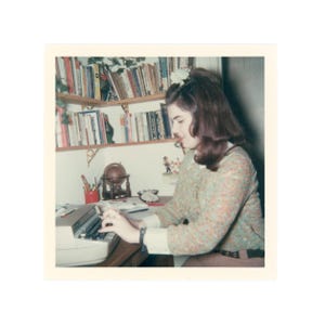 May include: Vintage photograph of a person typing on a typewriter at a desk. The person is wearing a floral patterned top and brown pants. A bookshelf filled with books is in the background, along with a small globe and other desk accessories.