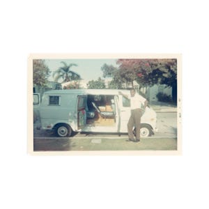 May include: Vintage photograph of a white van with the side door open, revealing the interior. A person in a white shirt and brown pants leans against the van. The photo is taken outdoors on a sunny day.