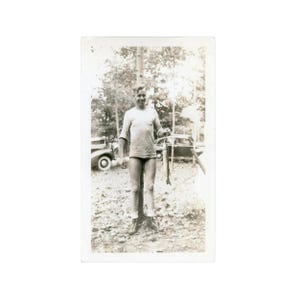 May include: A vintage black and white photograph showing a person holding two fish. The person is wearing a long-sleeved top, shorts, and boots. Cars and trees are visible in the background, suggesting an outdoor setting.