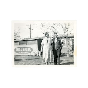 May include: Black and white photograph of a couple standing in front of a building with a sign that reads "Holsun Bread". The woman is wearing a dress, and the man is wearing a jacket and pants. The photo appears to be taken outdoors.