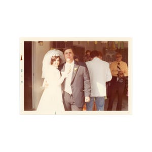 May include: Vintage photograph of a wedding couple. The bride wears a white wedding dress and veil, and the groom is in a dark suit with a light-colored vest. Other guests are visible in the background.