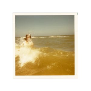 May include: A vintage square-format photograph showing a person partially submerged in ocean water. A wave is breaking in the foreground, creating white sea foam. The sky is clear and blue, and the overall color tone of the image is warm and sepia-tinted.