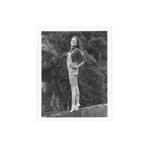 May include: Black and white photograph of a person wearing denim overalls and a long-sleeved shirt, standing on a wooden fence. The person is smiling, with hands on hips, and the background is a dense, dark green bush.