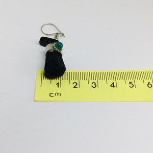 May include: A black and green dangle earring with a silver hook and a green stone. The earring is measured against a ruler that shows 1 cm.
