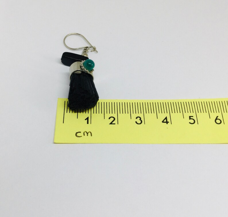 May include: A black and green dangle earring with a silver hook and a green stone. The earring is measured against a ruler that shows 1 cm.