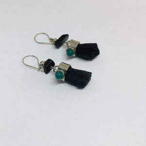 May include: A pair of earrings with black tassel-like charms and a green gemstone accent. The earrings have silver hooks and are made with black and silver metal.