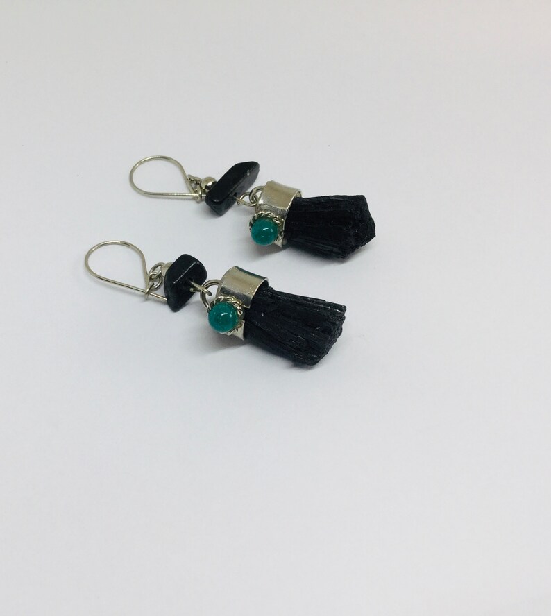 May include: A pair of earrings with black tassel-like charms and a green gemstone accent. The earrings have silver hooks and are made with black and silver metal.