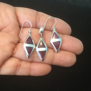 May include: A set of silver earrings and a pendant featuring a dark red gemstone in a diamond shape.