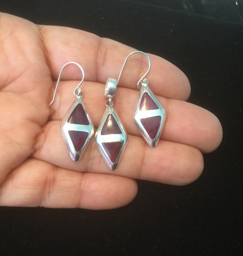 May include: A set of silver earrings and a pendant featuring a dark red gemstone in a diamond shape.