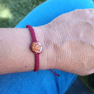 Può includere: Un braccialetto di corda rosso con un ciondolo rotondo marrone e dorato.