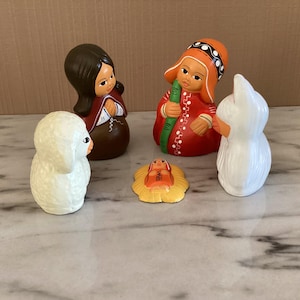 May include: A ceramic nativity set featuring five figures. The figures include a woman in a red and brown robe, a person in an orange hat and red robe, a white sheep, a baby in a yellow manger, and a woman in a red robe.