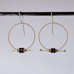 May include: A pair of hoop earrings crafted from coiled gold-toned wire. Each earring features a circular frame with a horizontal bar at the base, accented by two small black cylindrical beads and metallic silver-toned end caps on the wire.