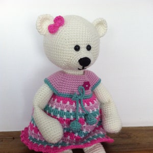 May include: A white crocheted teddy bear wearing a pink, green, and white dress with a pink bow on its head. The bear is sitting on a wooden surface.