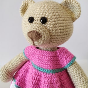 May include: A crocheted teddy bear wearing a pink dress with a green trim. The bear has brown button eyes and a brown nose.