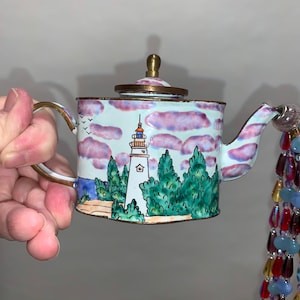 May include: A small white teapot with a brass lid and handle. The teapot is decorated with a painted scene of a lighthouse, trees, and clouds. The teapot is on a white background.