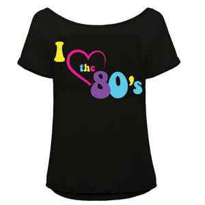 K&ouml;nnte beinhalten: Schwarzes Off-Shoulder-T-Shirt mit dem Schriftzug "I love the 80's" in Gelb, Rosa, Lila und Blau. Das Herz ist rosa und der Text in einer Retro-Schriftart gehalten, die an den Stil der 1980er erinnert.