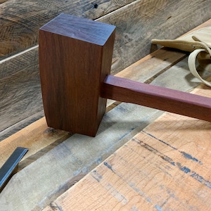 May include: A wooden mallet with a dark brown finish. The mallet has a rectangular head and a long, cylindrical handle.