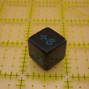 May include: A black six-sided die featuring teal numerical markings on its faces. The visible sides display the numbers plus six, plus two, and plus four in a simple font. The die has rounded corners and a smooth, solid finish.