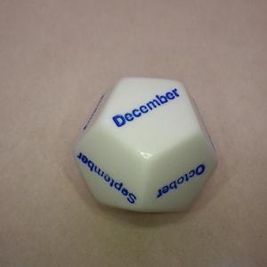 Pode incluir: Dado branco de seis lados com letras azuis. As faces do dado mostram os meses de setembro, outubro e dezembro.