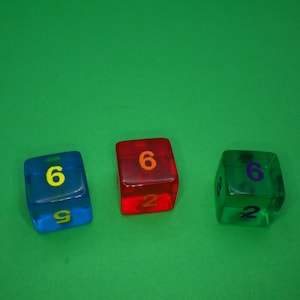 May include: Three clear dice with different colored plastic. The dice are numbered 6, 6, and 2. The dice are on a green surface.