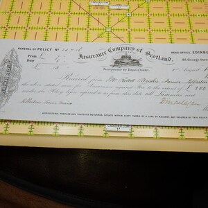 May include: An antique insurance receipt from the Insurance Company of Scotland, dated August 1, 1851. The document is on aged paper, with black ink text and decorative elements. It rests on a yellow and green grid ruler.