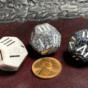 May include: Three speckled dice with white markings. The dice are a light gray, dark gray, and white. The dice are on a brown surface.
