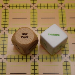 May include: Two dice with text on a yellow grid. One die is wooden with "HALF PLATE" printed on it. The other die is white with "CHAINMAIL" printed on it. The dice are used for crafting or gaming.