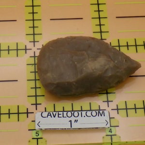 May include: A gray-brown, teardrop-shaped stone tool, approximately 1 inch in length, with a textured surface. The tool is displayed on a yellow and black grid background, with the text "CAVELOOT.COM" visible.