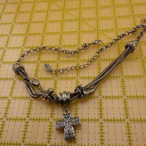 May include: A silver-toned necklace featuring a central cross pendant with intricate scrollwork and small clear stones. The chain consists of multi-strand flexible metal coils, decorative beads, and a standard link chain with a lobster claw clasp.