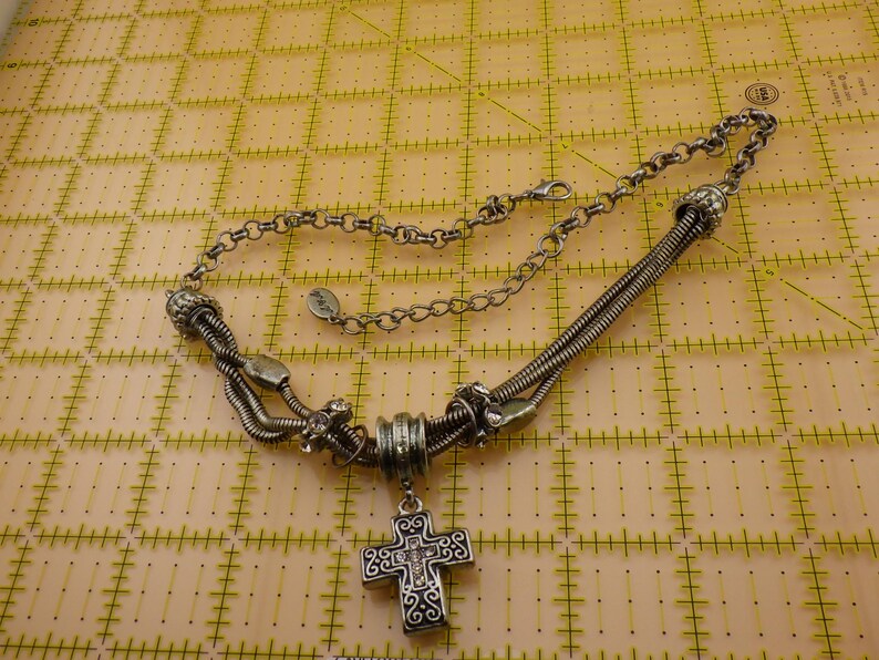 May include: A silver-toned necklace featuring a central cross pendant with intricate scrollwork and small clear stones. The chain consists of multi-strand flexible metal coils, decorative beads, and a standard link chain with a lobster claw clasp.