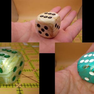May include: Three dice with different color schemes are shown. One is tan with black dots, another is green and white, and the third is teal with white dots. The dice are held in a hand, showcasing their size and design.