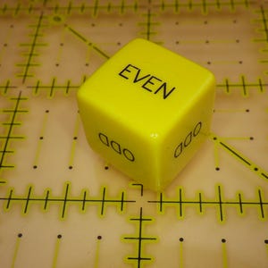 May include: A bright yellow six-sided die featuring black text on each face. The top face displays the word EVEN in capital letters, while the visible side faces display the word ODD in capital letters oriented vertically along the side.