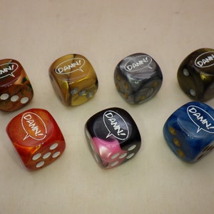 May include: Seven dice with the word "DAMN!" in a speech bubble on each side. The dice are in various colors including red, orange, yellow, black, blue, and gray.