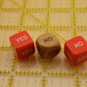 May include: Two red six-sided dice and one wooden six-sided die. The red dice feature the word YES in white outline font on multiple faces. The wooden die features the word NO in black outline font on the top face and YES on the side face.