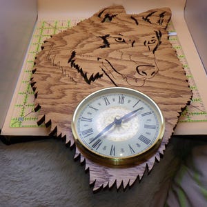 May include: A wooden clock featuring a carved wolf head design. The clock face is round with Roman numerals and a gold-colored bezel. The wood has a natural grain and the clock is approximately 12 inches wide.