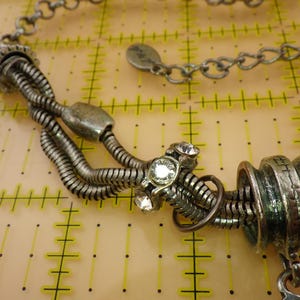 May include: A silver-toned necklace featuring multiple flexible snake chains, metallic cylindrical beads, and clear round rhinestones. The necklace includes a chain link extension with a small circular tag and is displayed over a yellow grid mat.