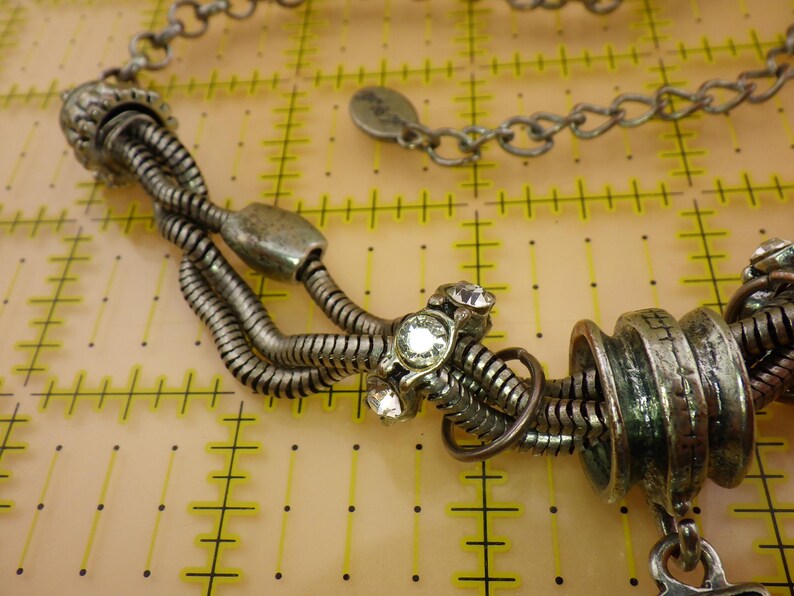 May include: A silver-toned necklace featuring multiple flexible snake chains, metallic cylindrical beads, and clear round rhinestones. The necklace includes a chain link extension with a small circular tag and is displayed over a yellow grid mat.