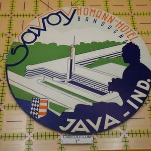 May include: A circular vintage-style sign featuring a graphic illustration of the Savoy Homann-Hotel in Bandoeng, Java, Indonesia. The design includes the text Savoy Homann-Hotel Bandoeng and Java-Ind. A 1-inch measuring tool is visible at the bottom.