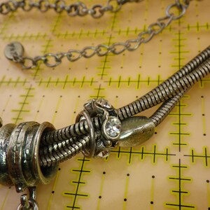 May include: A silver-toned necklace featuring a multi-strand snake chain design with metallic beads and clear rhinestones. The necklace includes a circular tag with the text 'PISA' and is positioned over a yellow grid mat with 1-inch markings.