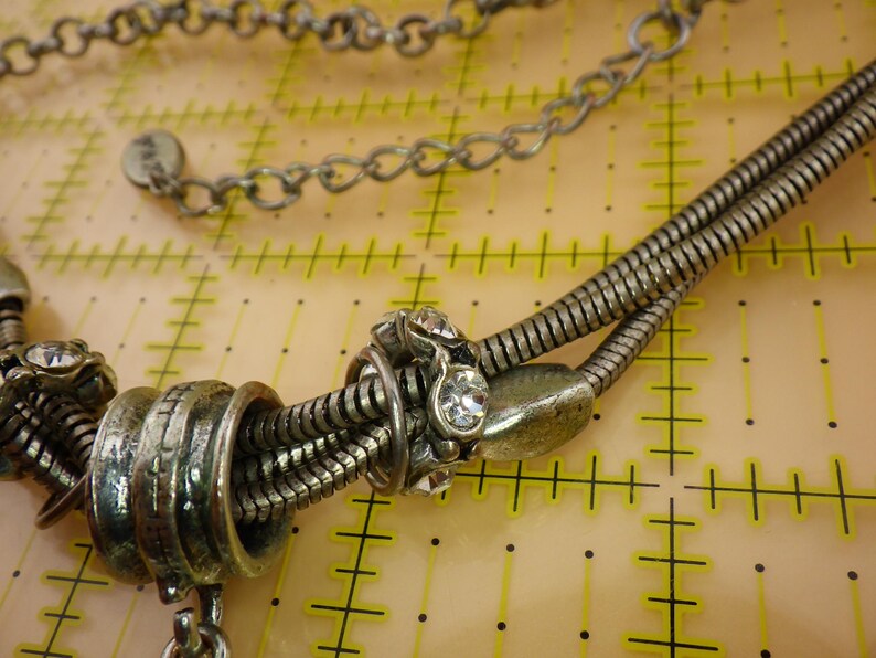 May include: A silver-toned necklace featuring a multi-strand snake chain design with metallic beads and clear rhinestones. The necklace includes a circular tag with the text 'PISA' and is positioned over a yellow grid mat with 1-inch markings.