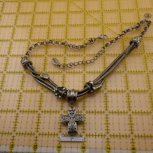 May include: A silver-toned necklace featuring a cross pendant with intricate scrollwork and small embedded stones. The necklace includes a multi-strand snake chain design with decorative metal beads and a lobster clasp. A 1-inch scale is visible.