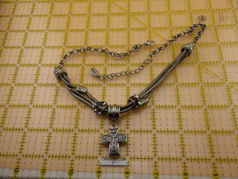 May include: A silver-toned necklace featuring a cross pendant with intricate scrollwork and small embedded stones. The necklace includes a multi-strand snake chain design with decorative metal beads and a lobster clasp. A 1-inch scale is visible.